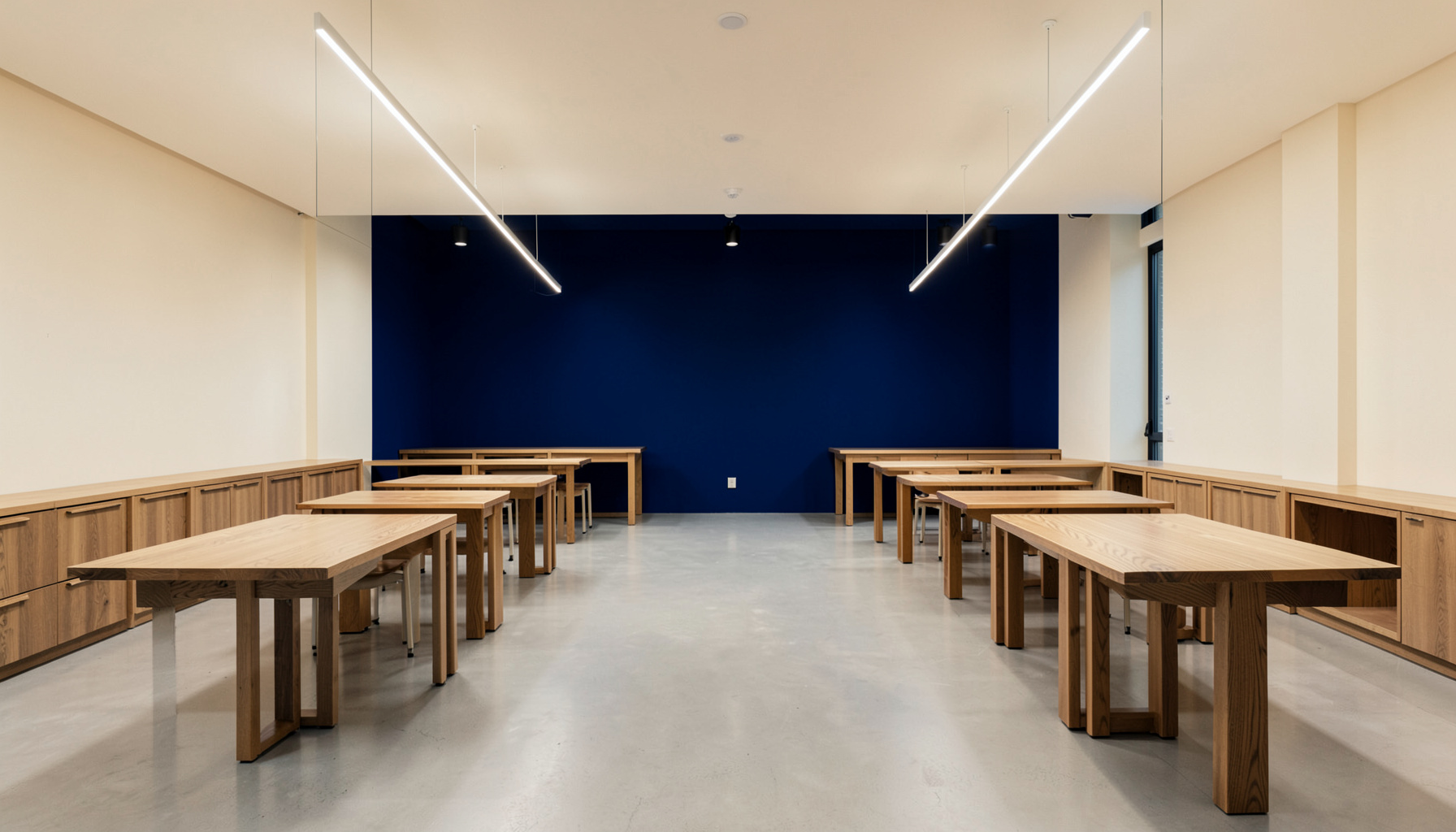 A clean architectural photograph with strong symmetry, showing a modern open-plan workspace with warm wood tables and soft overhead lighting in neutral cream and navy tones